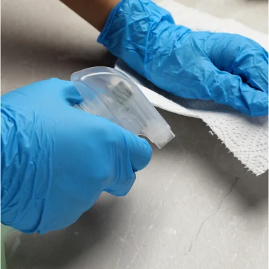 Mãos com luvas azuis borrifando lÃquido em um pano de papel branco.
