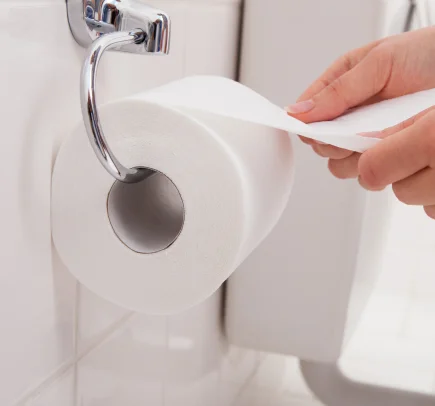 Mãos puxando papel higiênico branco de um suporte de metal fixado na parede de um banheiro.