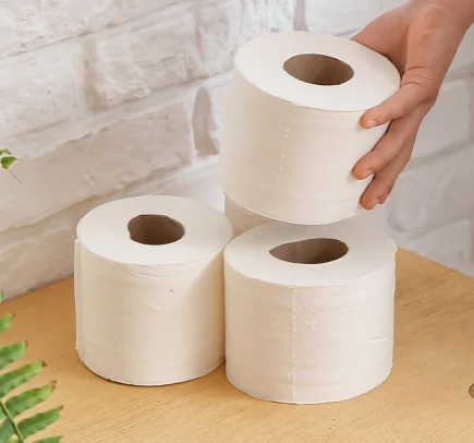 Três rolos brancos de papel higiênico empilhados sobre uma superfície de madeira, com uma mão segurando o rolo superior.