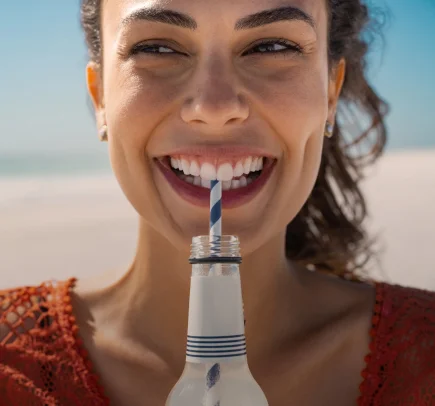 Mulher sorridente bebendo com canudo de uma garrafa branca na praia com céu azul ao fundo.