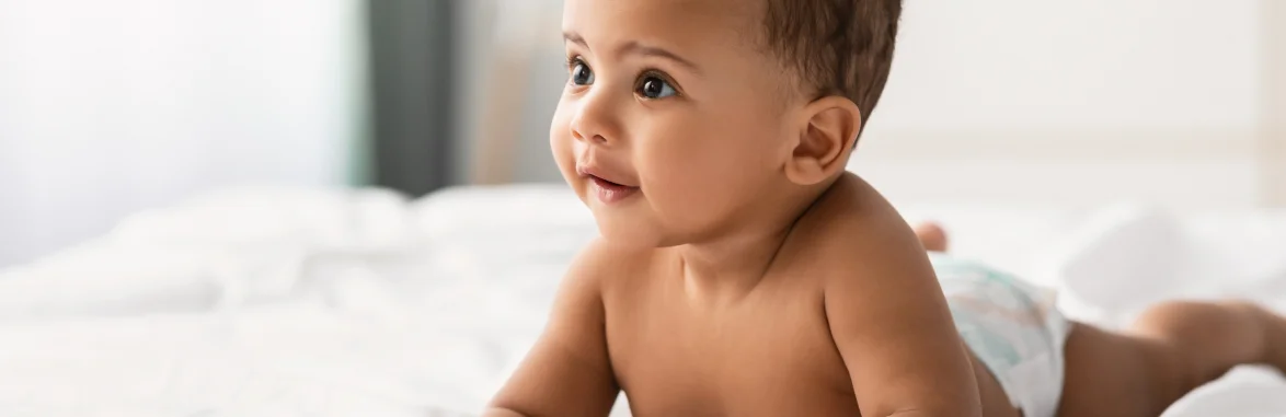 Bebê sorridente deitado de barriga para baixo sobre uma cama com lençol branco.