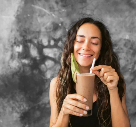 Mulher sorridente com olhos fechados segurando um copo de smoothie castanho com decoração verde e um canudo branco.