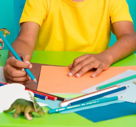 Criança usando camiseta amarela segurando lápis azul sobre folhas coloridas em uma mesa verde, com brinquedos de dinossauros ao lado.