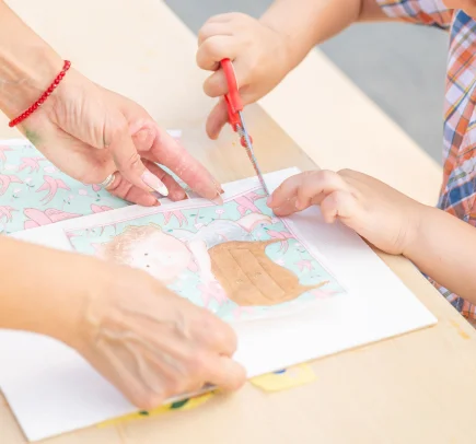 Mãos de uma criança cortando papel com tesoura vermelha com a ajuda de um adulto.