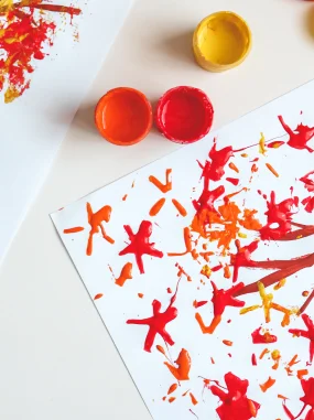 Pincel e tintas vermelha, laranja e amarela com desenhos coloridos em papel branco.