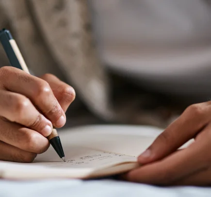 Mãos segurando uma caneta e escrevendo em um caderno aberto.