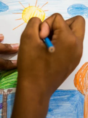 Mão segurando lápis azul enquanto desenha um sol amarelo em papel com céu azul, nuvens, árvore e grama.