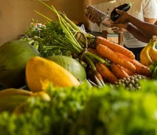 Pessoa escolhendo e pesando cenouras frescas em mercado com variedade de frutas e verduras.