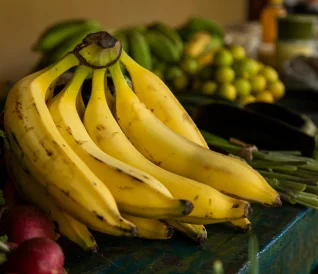 Cacho de bananas amarelas maduras em cima de uma mesa com outras frutas e legumes ao fundo.