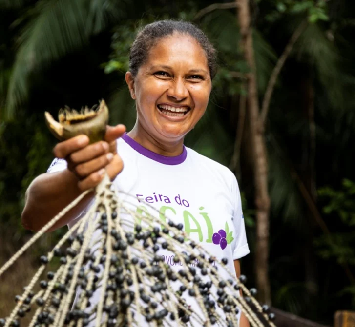 Mulher sorridente segura cachos de açaí em ambiente ao ar livre com vegetação ao fundo.