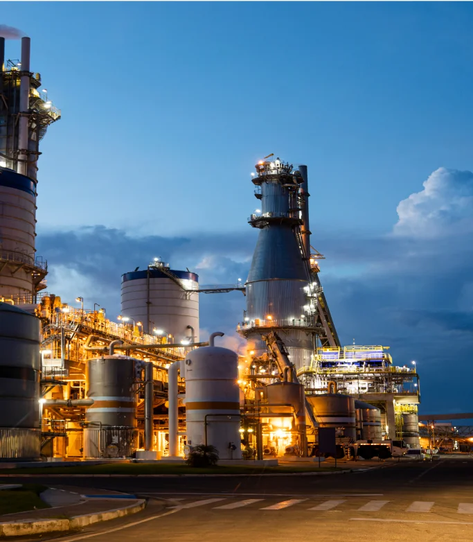 Instalação industrial iluminada à noite com tanques e chaminés contra um céu azul escuro.
