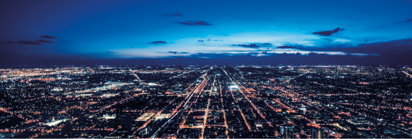 Vista panorâmica noturna de uma cidade com ruas iluminadas e céu azul escuro com poucas nuvens.