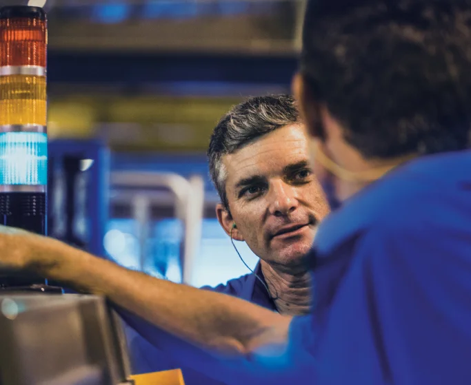 Dois homens em uniformes azuis conversando em uma fábrica, um deles está próximo a um painel com luzes indicadoras coloridas.