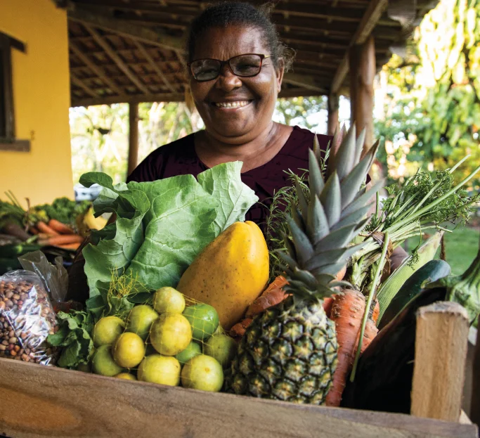 Mulher sorridente segurando caixa de madeira com frutas e verduras frescas, incluindo abacaxi, mamão, cenouras e folhas verdes.