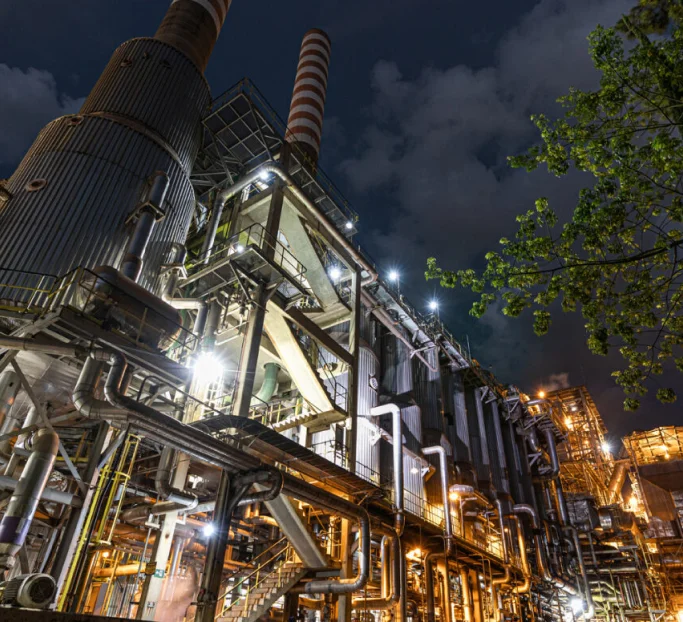 Planta industrial iluminada à noite com chaminés altas e estruturas metálicas complexas sob céu escuro com algumas nuvens.