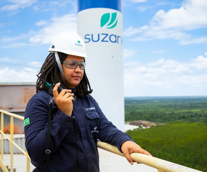 Mulher usando capacete branco e óculos de proteção em área industrial com microfone de rádio comunicador.