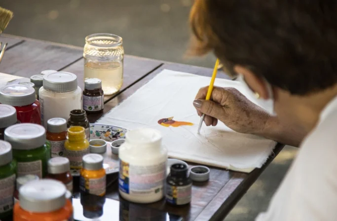 Pessoa pintando um peixe colorido em uma tela branca usando um pincel amarelo, com vários potes de tinta sobre a mesa de madeira.