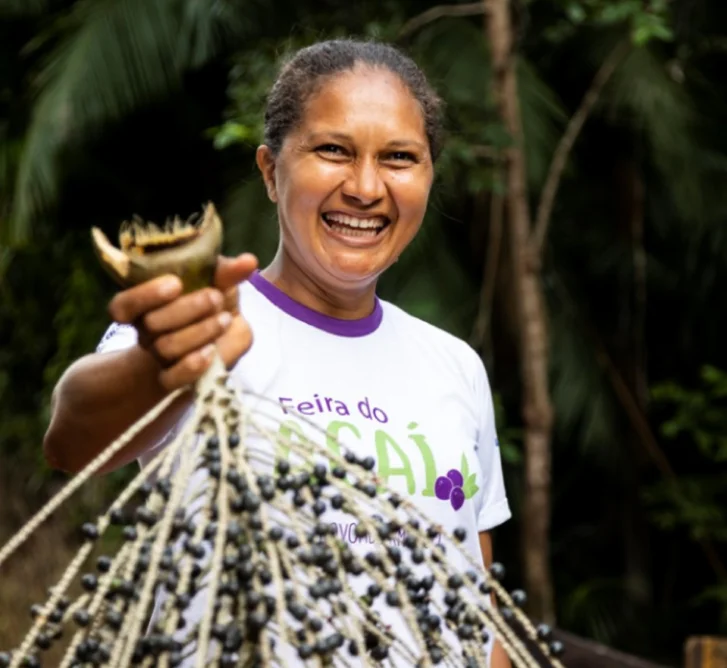 Mulher sorridente segurando um cacho grande de açaí em um ambiente ao ar livre com vegetação ao fundo.