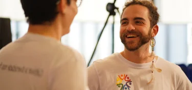 Duas pessoas sorrindo e conversando, uma usa uma camiseta branca com um logotipo colorido representando diversidade.