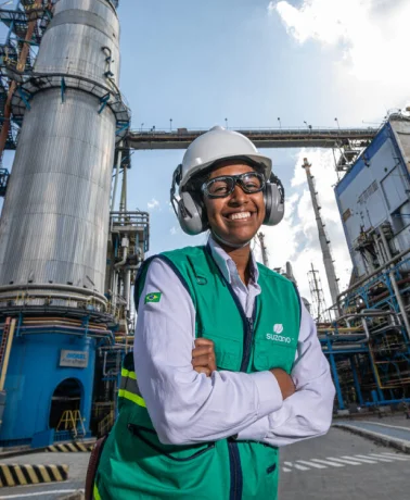 Trabalhadora sorridente usando capacete, protetores auriculares e colete verde em uma planta industrial.