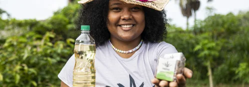 Mulher sorridente com chapéu segurando uma garrafa plástica e uma caixa pequena ao ar livre numa área verde.