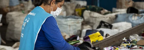 Mulher usando máscara e colete azul trabalha separando materiais recicláveis em linha de triagem.