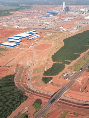 Vista aérea de um complexo industrial com várias construções de teto azul cercado por áreas de terra vermelha e plantações verdes.