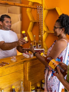 Homem atendendo cliente mulher em loja amarela com prateleiras hexagonais, entregando produto em frasco.