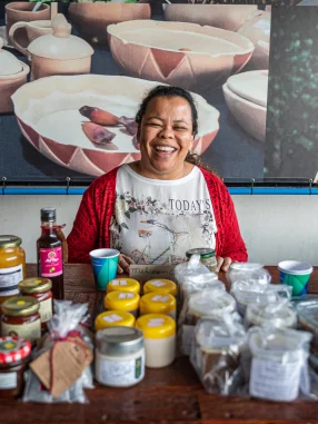 Mulher sorridente sentada atrás de mesa com potes e frascos de produtos artesanais em São Paulo.