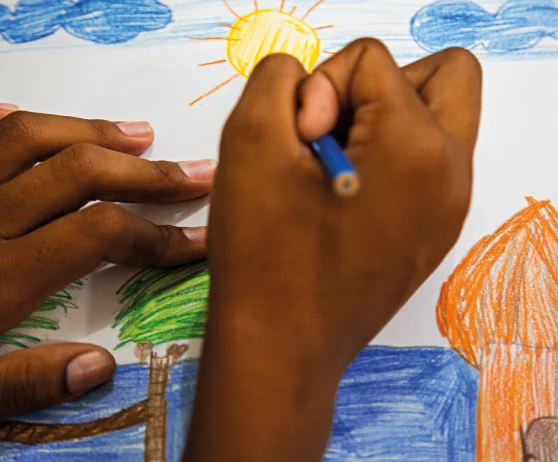 Mãos desenhando com lápis de cor uma paisagem com céu azul, sol amarelo, árvores verdes e uma casa laranja.