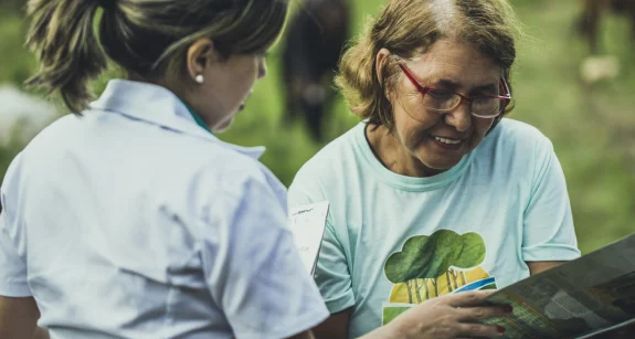 Duas mulheres conversam, uma delas usa jaleco branco e a outra, camiseta clara com estampa, segurando um mapa ao ar livre.