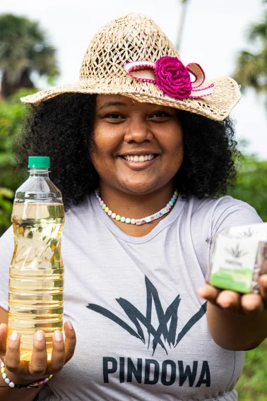 Mulher sorridente com chapéu de palha segurando uma garrafa plástica com líquido amarelo em uma mão e uma caixa pequena na outra, vestindo camiseta com logo Pindowa.