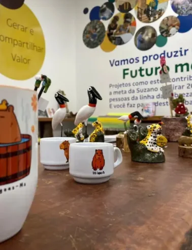 Canecas brancas com desenhos de animais, incluindo araras e onças-pintadas, expostas sobre mesa de madeira.