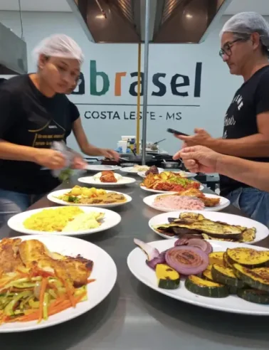 Duas pessoas com toucas servem pratos variados de comida em uma mesa com o logo da Abrasel Costa Leste - MS ao fundo.