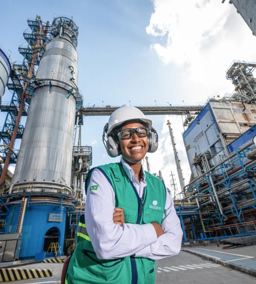 Funcionária sorridente com capacete, colete verde e protetores auriculares posando em frente a uma planta industrial de grande porte.