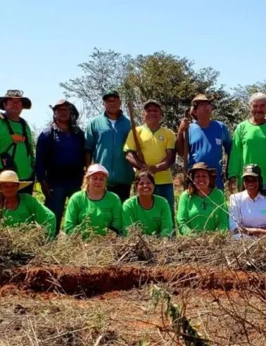 Grupo de pessoas vestindo camisas verdes e coloridas posando ao ar livre em área rural com vegetação seca e árvores ao fundo.