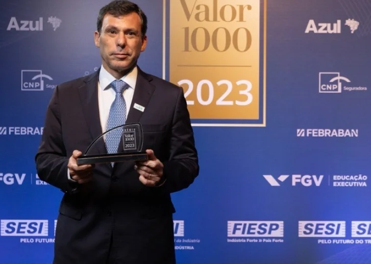 Homem de terno segurando um troféu do prêmio Valor 1000 2023, com fundo azul contendo logotipos de Azul, CNP Seguradora, Febraban, FGV, SESI e FIESP.