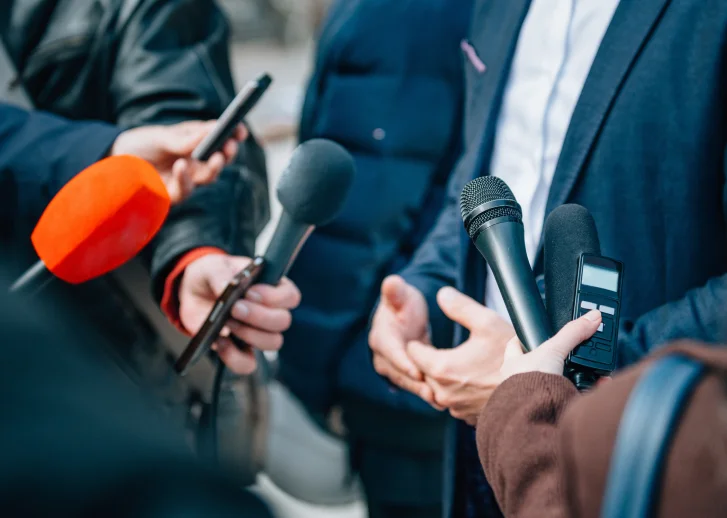 Pessoas segurando microfones e gravadores durante uma entrevista com um homem de terno azul.