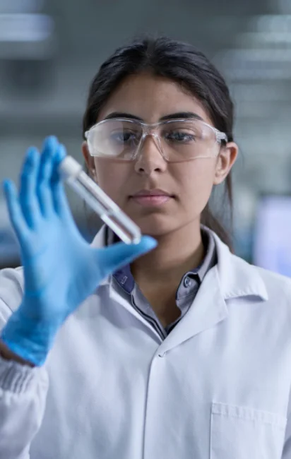 Cientista mulher vestindo jaleco branco, luvas azuis e óculos de proteção, segurando um tubo de ensaio transparente em laboratório.