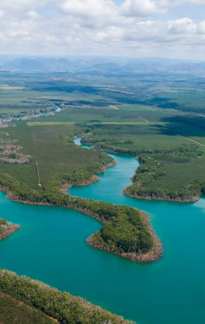 Vista aérea de um rio sinuoso com água azul-turquesa cercado por vegetação densa e florestas, com montanhas ao fundo sob céu nublado.