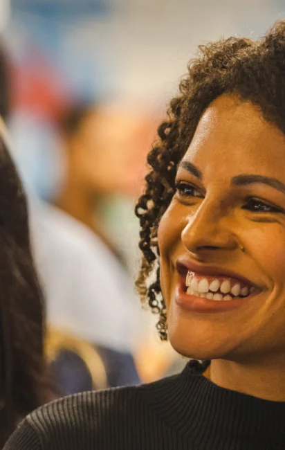 Close-up de uma mulher sorridente com cabelo cacheado e roupa preta, com fundo desfocado.