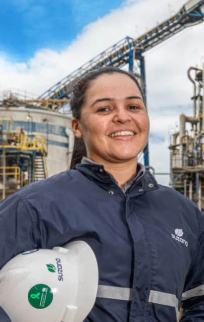 Mulher sorridente vestindo uniforme azul escuro da �ڰ������� segura capacete branco em uma fábrica ao ar livre.