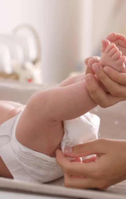 Mãos de adulto vestindo uma fralda em um bebê deitado, com fundo desfocado em tom neutro.