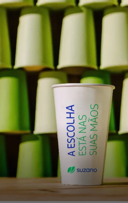 Copo branco descartável com texto em azul e verde dizendo 'A ESCOLHA ESTÁ NAS SUAS MÃOS' com o logo da Suzano, em frente a uma parede com copos verdes empilhados.