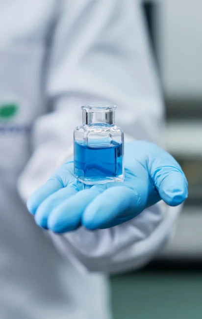 Mão com luva azul segurando um frasco de vidro com líquido azul em laboratório.