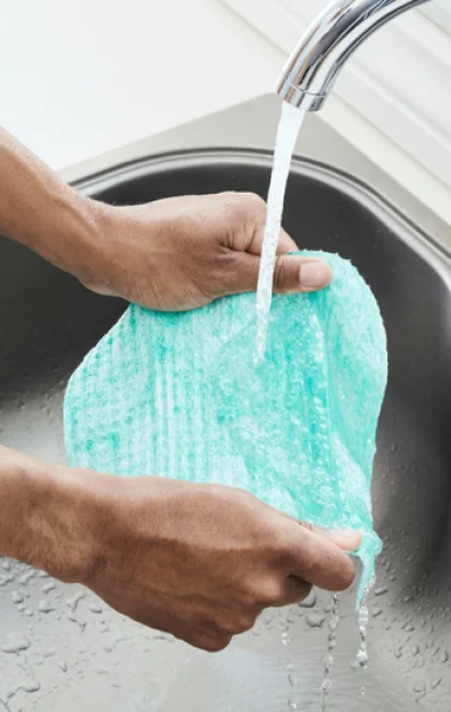 Mãos lavando pano reutilizável azul sob torneira com água corrente em pia.