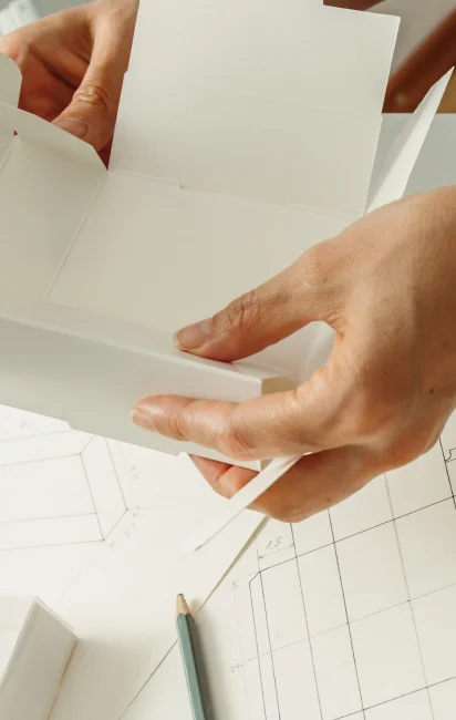 Mãos segurando uma caixa de papel cartão branca montada sobre uma mesa com desenhos técnicos e um lápis.