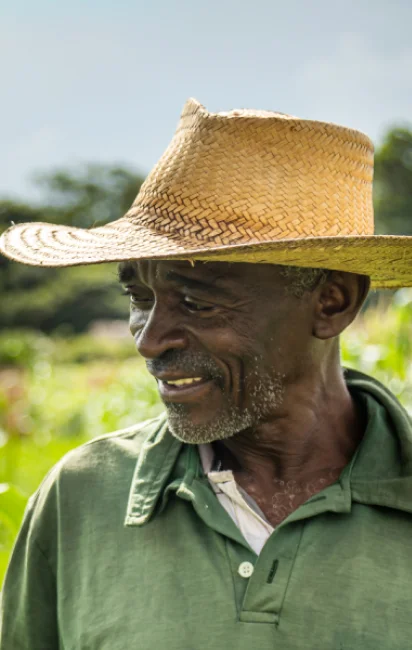 Homem idoso usando chapéu de palha e camisa verde sorrindo em um ambiente ao ar livre com vegetação ao fundo.