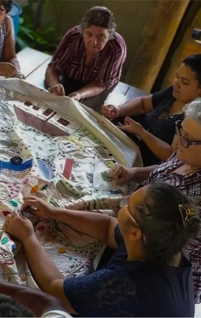 Grupo de mulheres bordando coletivamente um tecido grande com desenhos coloridos em uma mesa.