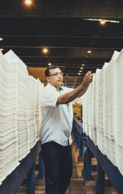 Homem inspecionando pilhas altas de folhas de papel empilhadas em um ambiente industrial.